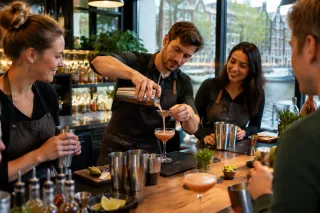 Man schenkt cocktail in coupeglas tijdens workshop, deelnemers lachen aan bar met ingrediënten